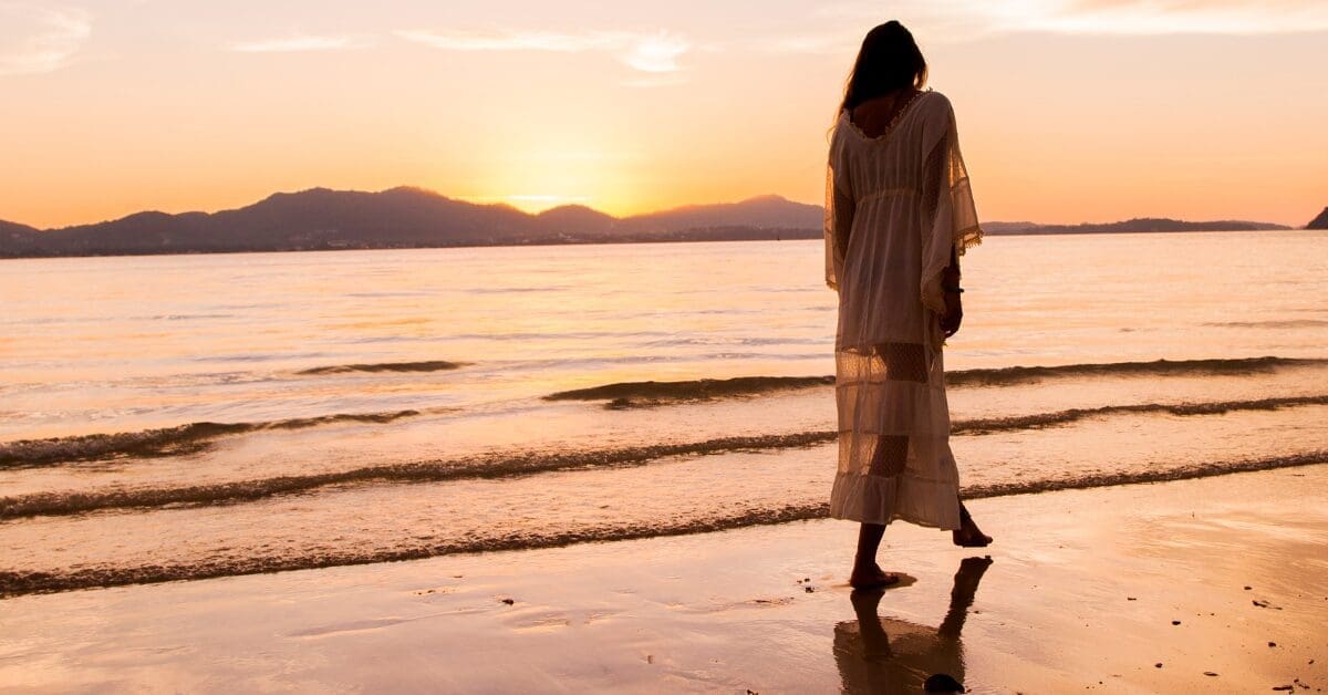 A woman in a flowing white dress walks barefoot along a wet sandy beach at golden sunset, her silhouette reflected in the shoreline as mountains rise softly in the distance — evoking the peace and solitude of a solo retreat experience.